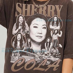 May include: A dark brown t-shirt featuring a large graphic print of Sherry Cola. The design includes multiple black and white photos of the person, a bus, a car, a microphone, and the text Sherry Cola. The shirt is labeled as Comfort Colors Espresso.
