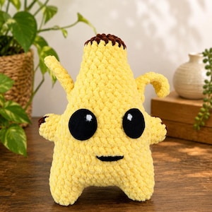 May include: A yellow crochet plush toy shaped like a banana with a brown top, two small arms, and two legs. The plush features large, round black eyes with white highlights and a small, horizontal black stitched mouth on its front surface.