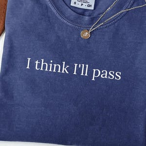 May include: A navy blue Comfort Colors t-shirt with the phrase "I think I'll pass" in white lettering. A gold necklace with a pendant rests on the shirt. A brown hat and dried plant are in the background.