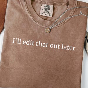 May include: A brown short-sleeved crew neck t-shirt with the white text I'll edit that out later printed on the front. The shirt features a Comfort Colors brand label at the inner neckline indicating a size small and 100% cotton material.