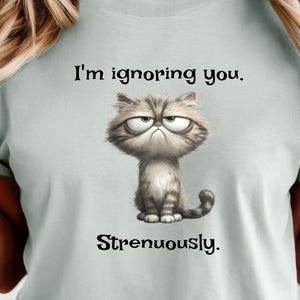 Könnte beinhalten: Hellgrünes T-Shirt mit einem mürrischen Cartoon-Kater und dem Text "I'm ignoring you. Strenuously." Die Katze ist grau und weiß mit einem strengen Gesichtsausdruck. Das T-Shirt hat kurze Ärmel und einen Rundhalsausschnitt.