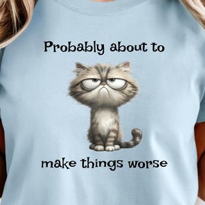 May include: Light blue t-shirt with the text "Probably about to make things worse" and a grumpy cartoon cat illustration. The cat is gray and white with a furrowed brow.
