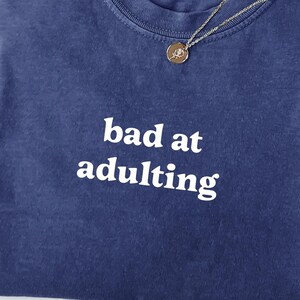 May include: A navy blue t-shirt with the phrase "bad at adulting" in white block letters. A gold necklace with a circular pendant rests on the shirt. A brown hat and dried plant are in the background.