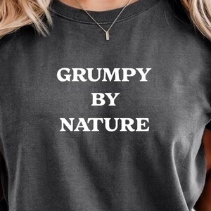 Op de afbeelding: Donkergrijs T-shirt met de witte tekst "GRUMPY BY NATURE". Het shirt heeft een ronde hals en is gemaakt van een zacht materiaal. Een zilveren ketting is zichtbaar. Een casual kledingstuk.