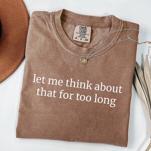 May include: A brown short-sleeved t-shirt with a crew neckline and the white text "let me think about that for too long" printed on the front. The shirt features a Comfort Colors brand label at the inner neck and is made of cotton fabric.