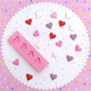 Può includere: Uno stampo in silicone rosa con cavità a forma di cuore, circondato da piccole caramelle a forma di cuore colorate in rosso, rosa, bianco e viola. Le caramelle sono sparse su una tovaglietta bianca con pois rosa.