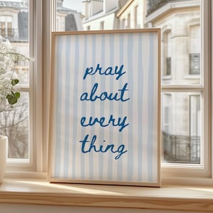 Könnte beinhalten: Ein gerahmter Druck mit den Worten "pray about everything" in blauer Schrift. Der Hintergrund zeigt vertikale hellblaue und weiße Streifen. Der Rahmen ist aus hellem Holz gefertigt, und der Druck wird auf einer Fensterbank ausgestellt.