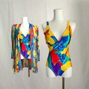 May include: A one-piece swimsuit and a matching long-sleeved cover-up, both featuring a vibrant, multi-coloured abstract print with prominent shades of yellow, blue, red, and purple, displayed on two separate white mannequin torsos.