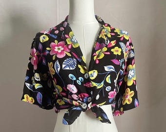 Könnte beinhalten: Schwarzes Oberteil mit floralem Print und Bindevorderseite. Das Oberteil hat ein lebendiges Muster aus rosa, gelben, blauen und weißen Blumen und Blättern. Die kurzärmlige Bluse wird auf einer Schaufensterpuppe präsentiert.