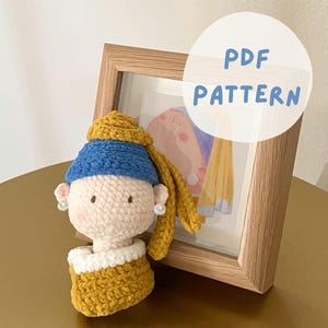 Puede incluir: Patrón de amigurumi de crochet de una niña con una diadema azul y cabello amarillo, que lleva un vestido amarillo. La muñeca está sentada frente a una imagen enmarcada con el texto "PDF PATTERN".