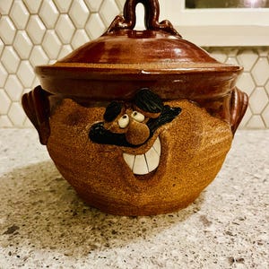 May include: A brown ceramic lidded pot with a smiling face design. The pot has a dark brown lid and handle, with a lighter brown body. The face features black hair, a mustache, and white teeth.