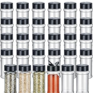 May include: A collection of clear spice jars with black lids. Some jars are filled with various spices, including black pepper, beige granules, green herbs, and red paprika. The jars are arranged in vertical rows.