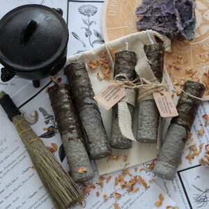 May include: A black cast iron cauldron, a small straw besom with a moon charm, and four cylindrical candles with a dark brown wax top and a textured grey base. The candles are wrapped in burlap ribbon with small paper tags and scattered dried petals.