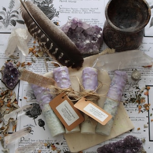 May include: Three cylindrical candles with light purple wax tops and textured white bases, each wrapped in burlap twine with a brown paper tag labelled Creator (God) Spirit kruidenkaars and Creator Wood Spirit kruidenkaars.