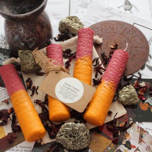 Puede incluir: Tres velas cilíndricas con una mitad superior rosa texturizada y una mitad inferior naranja lisa. Una etiqueta de papel marrón atada con cordel dice Hestia (Vesta) Kruidenkaars, rodeada de pétalos de flores secas y piedras de pirita.