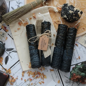 May include: Four textured black pillar candles with white wicks, arranged on a cream-coloured fabric pouch. One candle is tied with a hessian ribbon and a small brown paper tag labelled Seth kruidenkaars. The candles are surrounded by scattered dried petals.