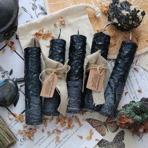 May include: Five black pillar candles with a textured, uneven surface. Two candles are wrapped in tan hessian ribbon with small brown paper tags labelled Hades Krudekaars. The candles are arranged near a small black cauldron, crystals, and dried petals.