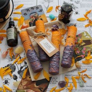 May include: Four cylindrical orange candles with purple wax drips at the base, tied with twine and a paper tag labeled Lugh (Lugos) kruidenkaars. The candles are arranged on a beige fabric pouch surrounded by yellow flower petals and crystals.