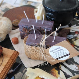 May include: Two rectangular purple candles with white wicks, one wrapped in burlap with a tan paper tag labelled Mid-Samhain Kruidenkaars Moon Candle Craft. The candles are surrounded by a fossil, a black cauldron, a wooden pentacle, and stones.