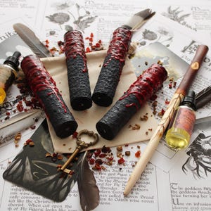 Puede incluir: Cuatro velas negras con superficies texturizadas y detalles de cera roja. Las velas están dispuestas con una varita de madera, una llave y una botella de aceite sobre un fondo de papel con texto y plumas. Tema místico.