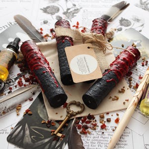 May include: Three black cylindrical candles with textured surfaces and red wax drips, tied with burlap ribbon and a round paper tag labeled Hekate Kruidenkaars. The candles are surrounded by feathers, a brass key, glass vials, and printed paper.