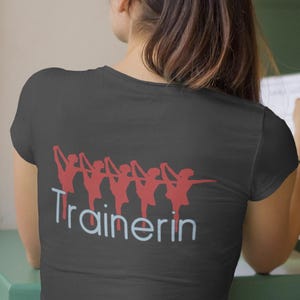 Könnte beinhalten: Dunkelgraues T-Shirt mit einer roten Grafik von Balletttänzerinnen und dem Wort "Trainerin" in Weiß und Hellblau. Das Shirt ist aus einem weichen Material.