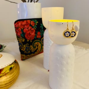 May include: White ceramic decorative vases with a face design, one with gold earrings. The earrings have a blue and white floral design. A vase holds branches. A patterned textile with red flowers is behind a vase.