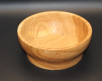 May include: A light-colored wooden bowl with a rounded shape and a pedestal base. The bowl has a smooth, polished finish, showcasing the natural wood grain. The bowl is empty and sits on a dark surface.
