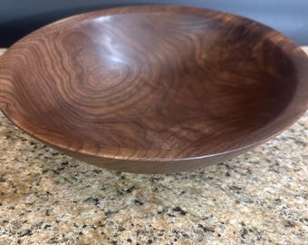 May include: A round wooden bowl featuring a dark brown color with visible natural wood grain patterns. The bowl has a smooth, polished finish and a wide, open interior, showcasing the organic texture and swirling lines of the wood material.