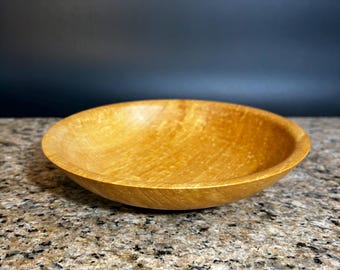 May include: A shallow, circular wooden bowl with a smooth, polished finish. The bowl features a warm, golden-brown color with visible natural wood grain patterns throughout the interior and exterior surfaces, showcasing a simple, rounded rim.