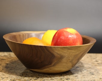 May include: A wooden bowl filled with fresh fruit. The bowl is made of a dark wood and contains a red apple, a yellow lemon, and an orange. The bowl sits on a speckled countertop with a dark background.