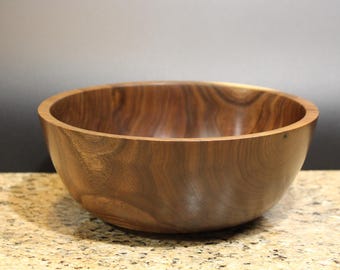 May include: A handcrafted wooden bowl with a rich, dark brown color. The bowl has a smooth, polished finish and a wide, open design. The wood grain is visible, adding to the natural aesthetic. This bowl is perfect for serving salads or displaying decorative items.
