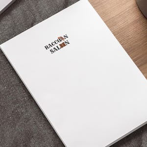 Könnte beinhalten: Ein weißer Notizblock mit dem Text "RACCOON SALOON" in brauner und roter Schrift. Der Notizblock liegt auf einer dunkelgrauen Oberfläche, mit einem Laptop und einem Stift im Hintergrund. Eine Topfpflanze befindet sich in der oberen rechten Ecke.