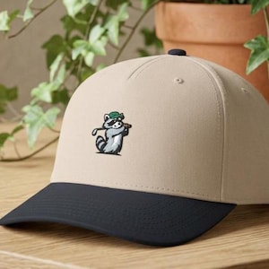 Peut inclure: Une casquette de baseball beige avec une visière bleu marine foncé. L'avant de la casquette présente un motif brodé représentant un raton laveur gris et blanc portant un chapeau vert et tenant un club de golf, en position de swing.