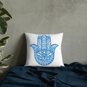 May include: White square pillow with a blue Hamsa hand design. The Hamsa hand features an eye in the center and intricate patterns. The pillow is placed on a dark blue blanket.