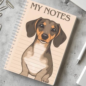 Puede incluir: Una libreta de espiral con una cubierta de color beige que presenta un dachshund de dibujos animados con pelaje marrón y beige. El texto "MY NOTES" está impreso en negro en la cubierta.