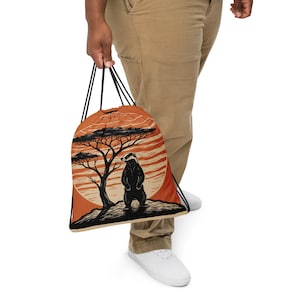 Puede incluir: Mochila con cordón naranja con un diseño de tejón en blanco y negro frente a un árbol y un atardecer. La bolsa tiene cordones negros y la sostiene una persona con pantalones color caqui y zapatos blancos.