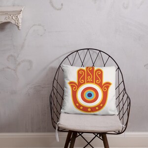 May include: A decorative cushion with a Hamsa hand design in orange, yellow, and red. The central eye motif is blue, white, and yellow. The cushion is placed on a metal chair. The chair is in front of a grey wall.