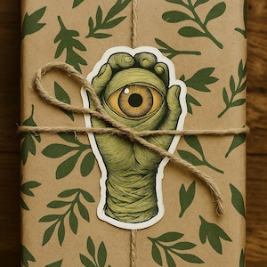 May include: A wrapped gift with a brown paper featuring green leaf patterns. A sticker of a green hand holding a yellow eye is centered on the package. The package is tied with twine.