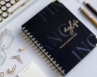 Può includere: Un diario nero a spirale con lettere dorate che recitano "FINANCING my life" e "BUDGET & GOALS PLANNER".