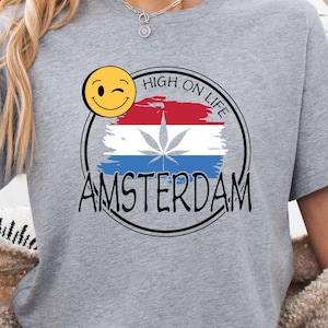 Może przedstawiać: Szara koszulka z nadrukiem przedstawiającym uśmiechniętą buźkę, napisem "HIGH ON LIFE" i "AMSTERDAM" w czarnym okręgu. Projekt zawiera czerwoną, białą i niebieską flagę z liściem konopi.