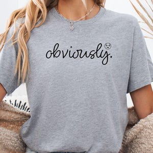 May include: A pale grey t-shirt featuring the word "obviously" in black cursive, with a small, sad-faced emoji. The shirt is worn with denim shorts and a cream cardigan.