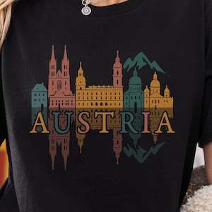 May include: Black t-shirt featuring a colorful graphic of the Austria skyline with a mirrored reflection. The word "AUSTRIA" is printed in a retro font, with a mountain range in the background.