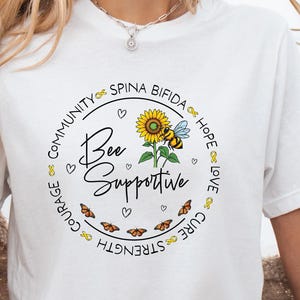 May include: White t-shirt with a black circle graphic featuring a sunflower, a bee, and the text "Bee Supportive". The circle is surrounded by yellow ribbons and the words "Community", "Courage", "Strength", "Cure", "Hope", and "Love".