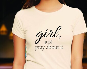 May include: Cream-colored t-shirt with the text "girl, just pray about it" in black script. The shirt has a crew neck and short sleeves. The text is centered on the front of the shirt.