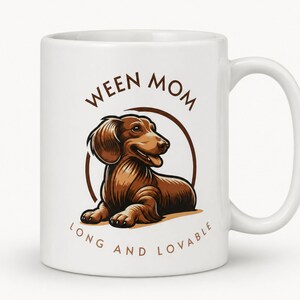 Può includere: Tazza in ceramica bianca con un'illustrazione di bassotto marrone all'interno di un cerchio marrone. Il testo "WEEN MOM" è arcuato sopra il cane, con "LONG AND LOVABLE" sotto. La tazza ha un manico bianco.