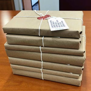 May include: A pile of brown paper-wrapped parcels, secured with white string. A red wax seal and a small tag are affixed to the top package. The parcels are stacked on a wooden surface.