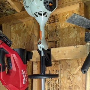 May include: A white and orange STIHL string trimmer is mounted on a wooden wall. A red Milwaukee leaf blower is visible on the left. The image is taken in a wooden shed.