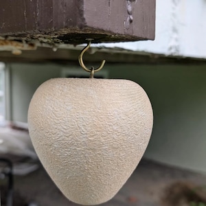 May include: A beige, heart-shaped decorative object with a textured surface, suspended from a brass hook. The hook is attached to a brown wooden beam. The background is blurred, suggesting an outdoor setting.