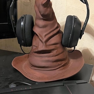 May include: A brown Sorting Hat figurine with a wide brim and a pointed top, with a pair of black headphones resting on it. The headphones have black earcups and a black headband. The hat is sitting on a black surface.